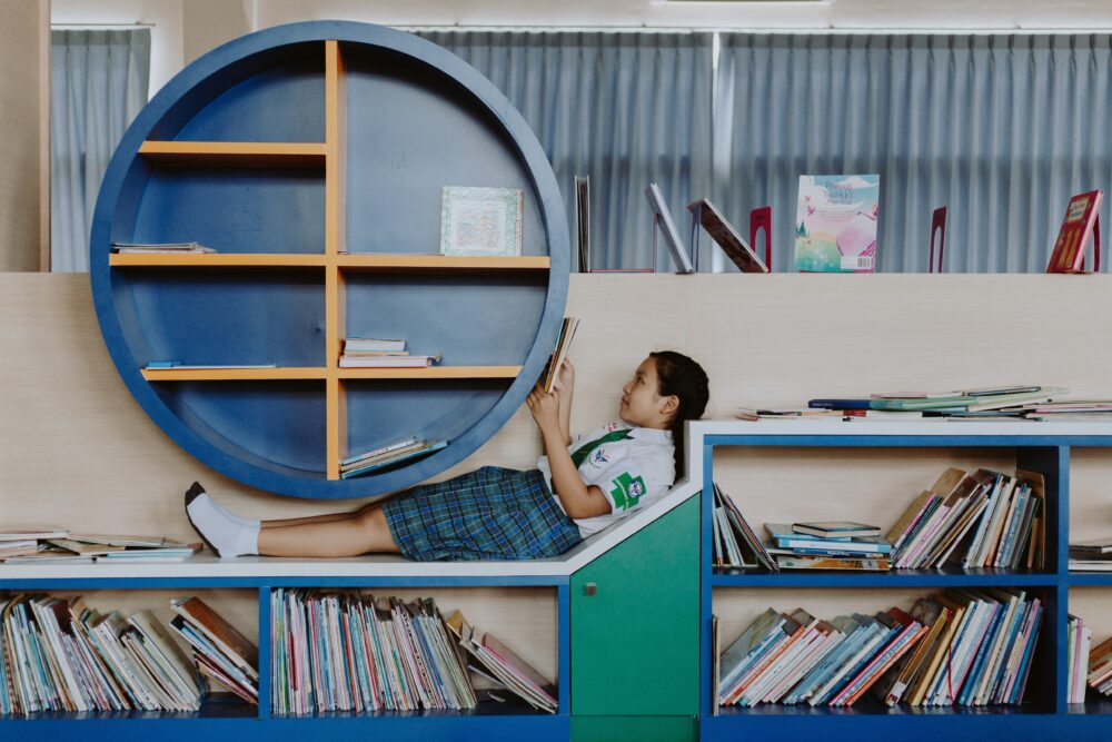Child reads a book in a library on top of a blue bookshelf filled with lots of book